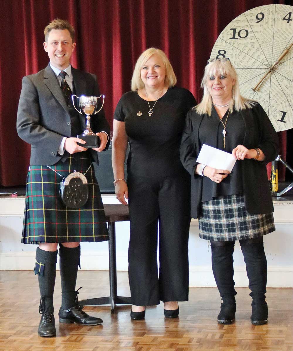 2023 City of Edinburgh Pipe Band Wheel of Fortune winner Cameron Drummond accepting the trophy from Debbie Reilly and Tracy Ann Miller.