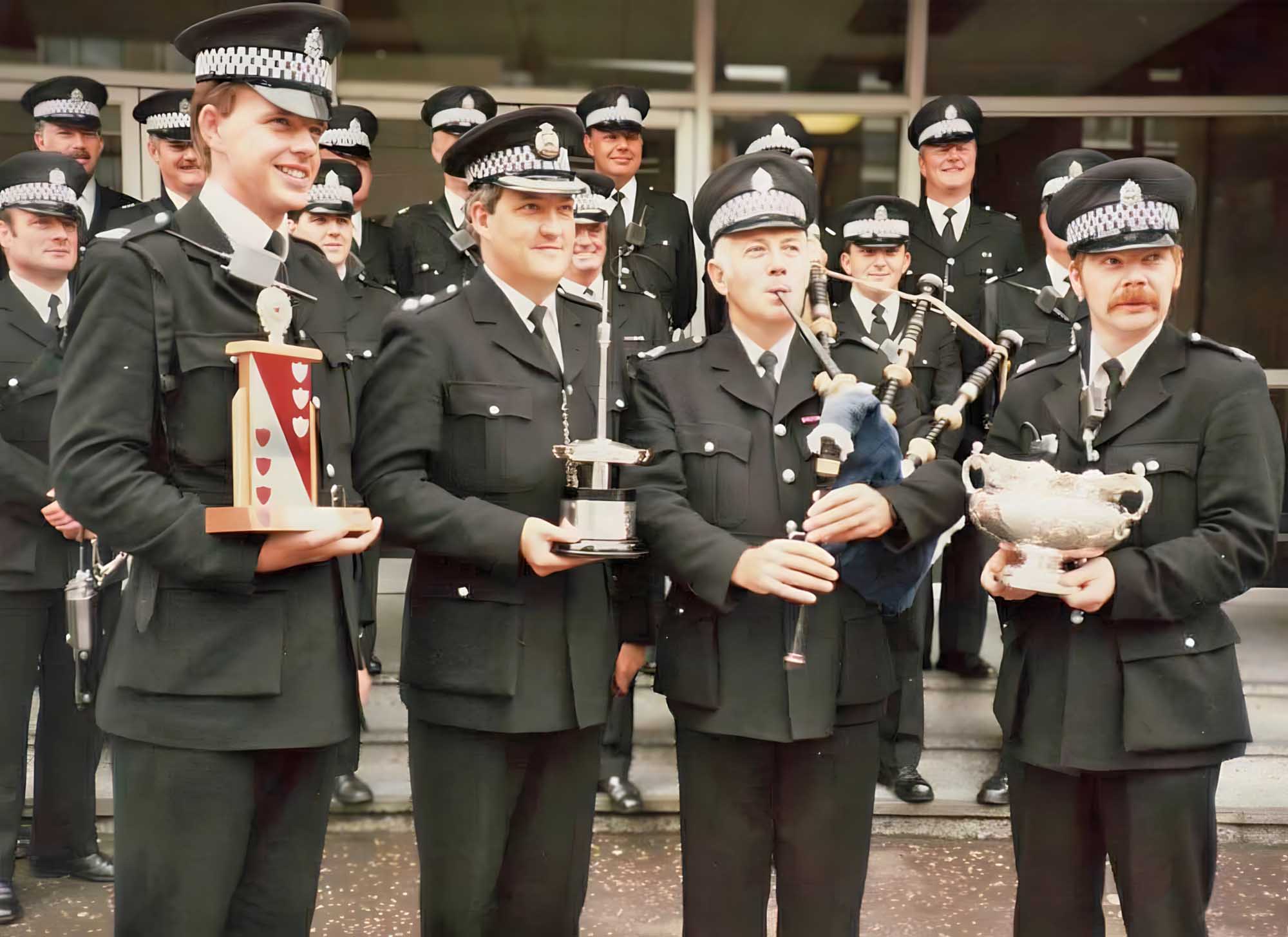 Strathclyde Police Pipe Band with the 1985 World Championship trophy. The front (L-R): Bass drummer Paul Harvey, John Wilson, Ian McLellan, and piper John Winter. Behind (L-R): James McLean, Jim Wark, Ian Kennedy, Wilson Brown, Harry McAleer, Bob Edmiston, Alex Connell, Ian Thomson, John Kirkwood and Graham Richardson. Behind Ian McLellan (L-R): Jim Semple, Jim McLuskey and Syd Spence.