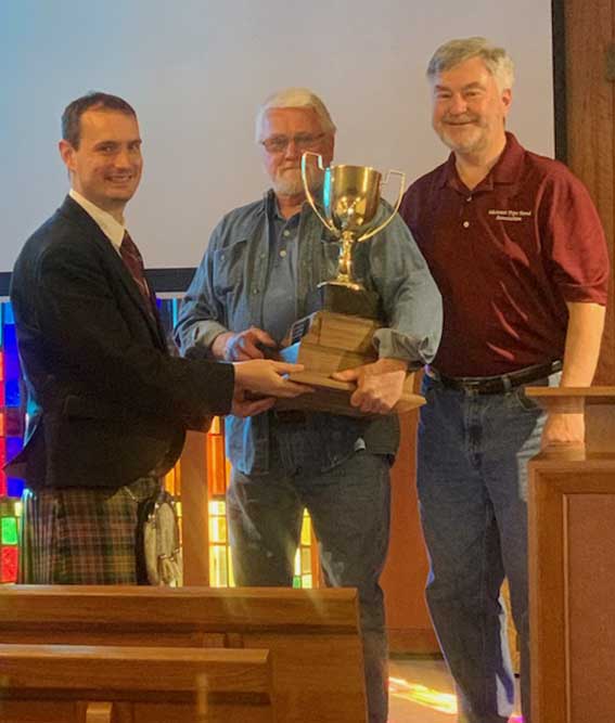 Andrew Carlisle (left) gets the trophy from Ian Swinton, namesake of the P-M Ian Swinton Open Piping Championship, with MWPBA President Jim Sim at right.